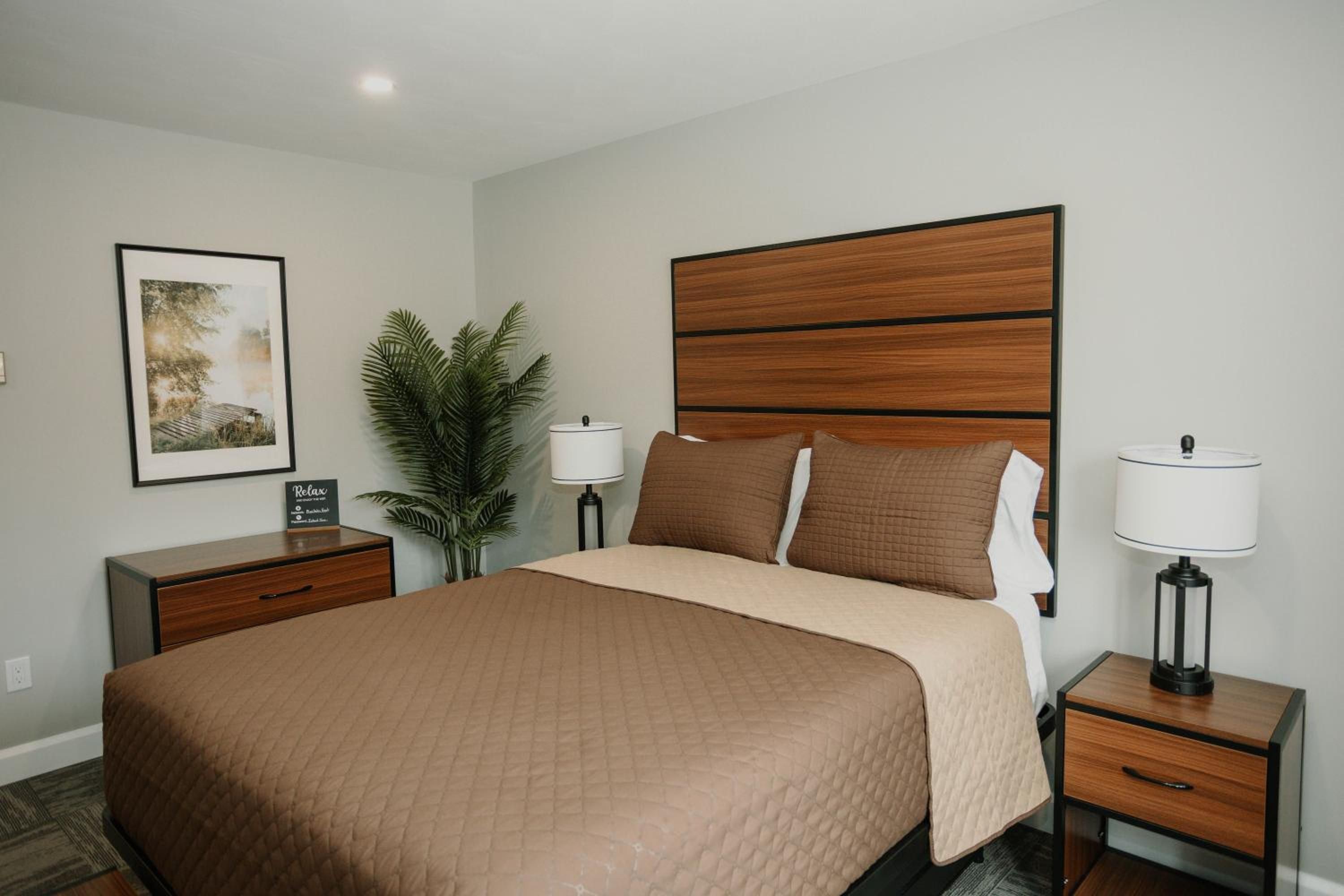 Une chambre d’hôtel avec un grand lit, deux tables de chevet, une petite table, un tableau encadré au mur et une plante en pot.
