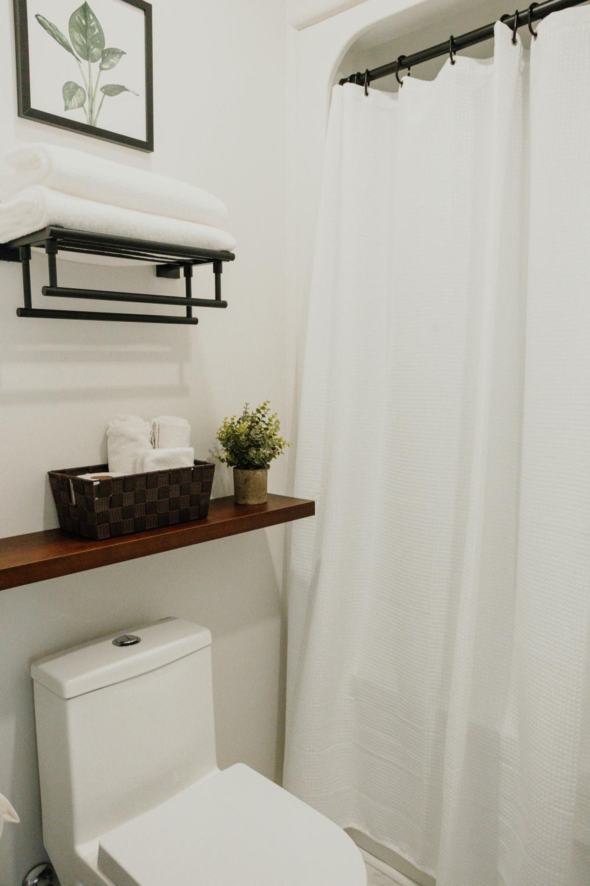 A bathroom with a white toilet, a shelf with a plant and a basket, and a white shower curtain.