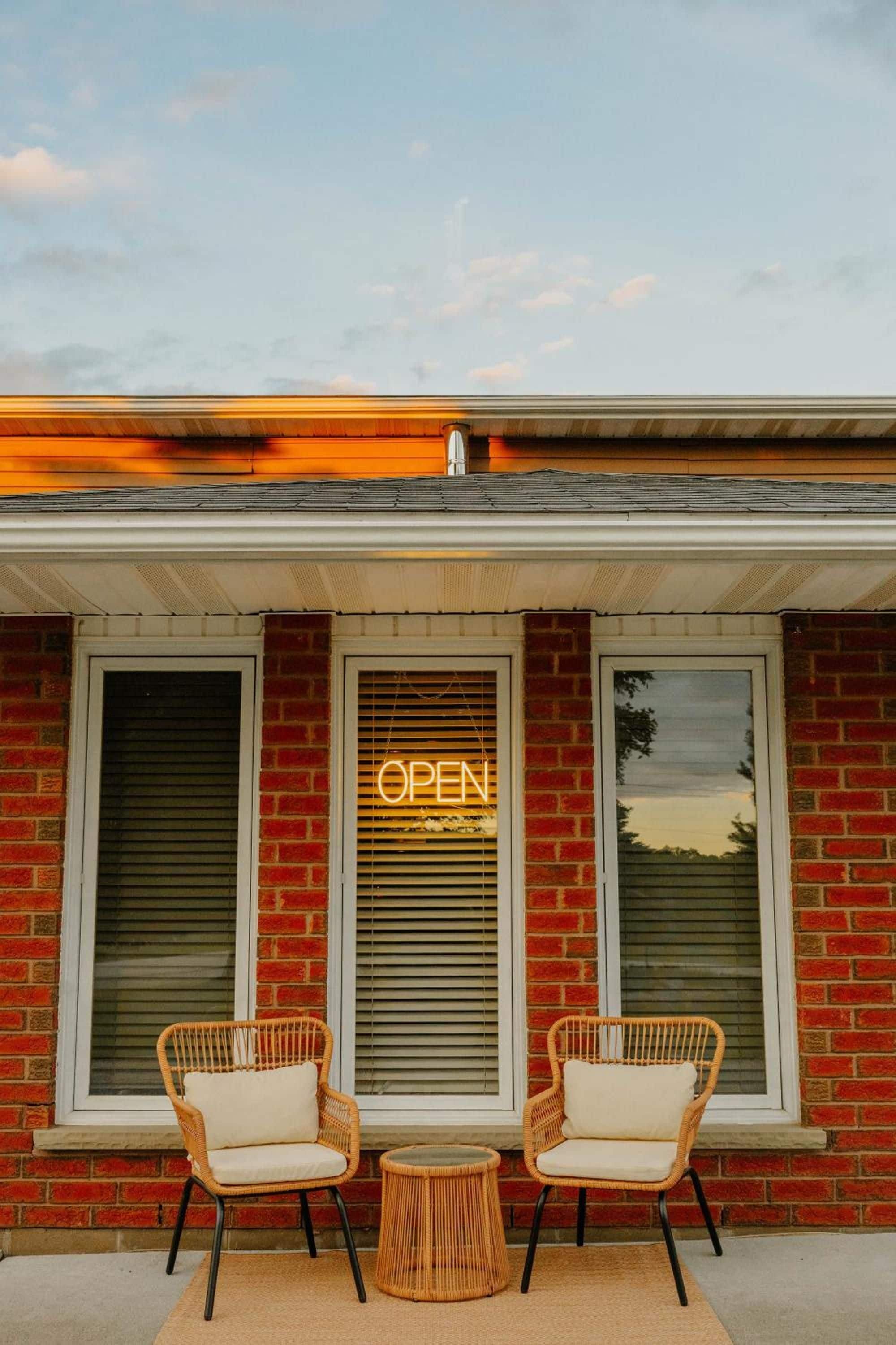 A building with a brick facade, two wicker chairs with cushions, and a neon 'OPEN' sign on the windows.