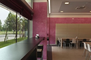 A modern interior with a long table and chairs, a window with a view of a road and trees, and a pink wall.