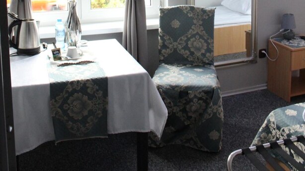 A hotel room with a bed, a chair, a table with a vase and a kettle, and a window with curtains. - Restauracja Hotel Kassandra (Września)