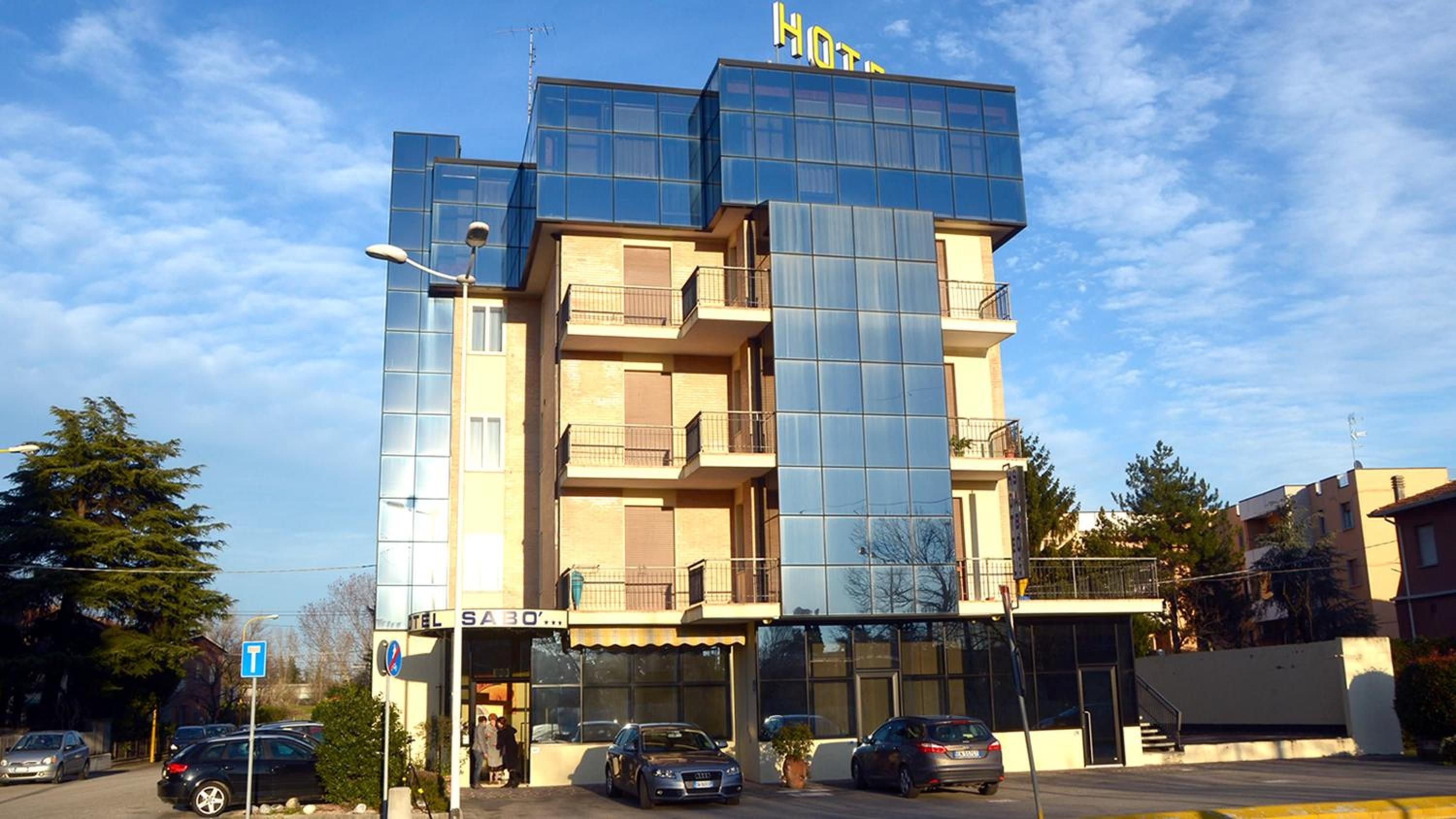 A multi-story hotel with a modern glass facade, a visible entrance, and a parking area with cars.