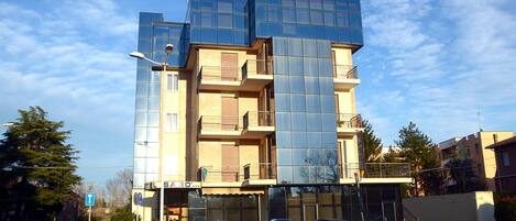 A multi-story hotel with a modern glass facade, a visible entrance, and a parking area with cars.