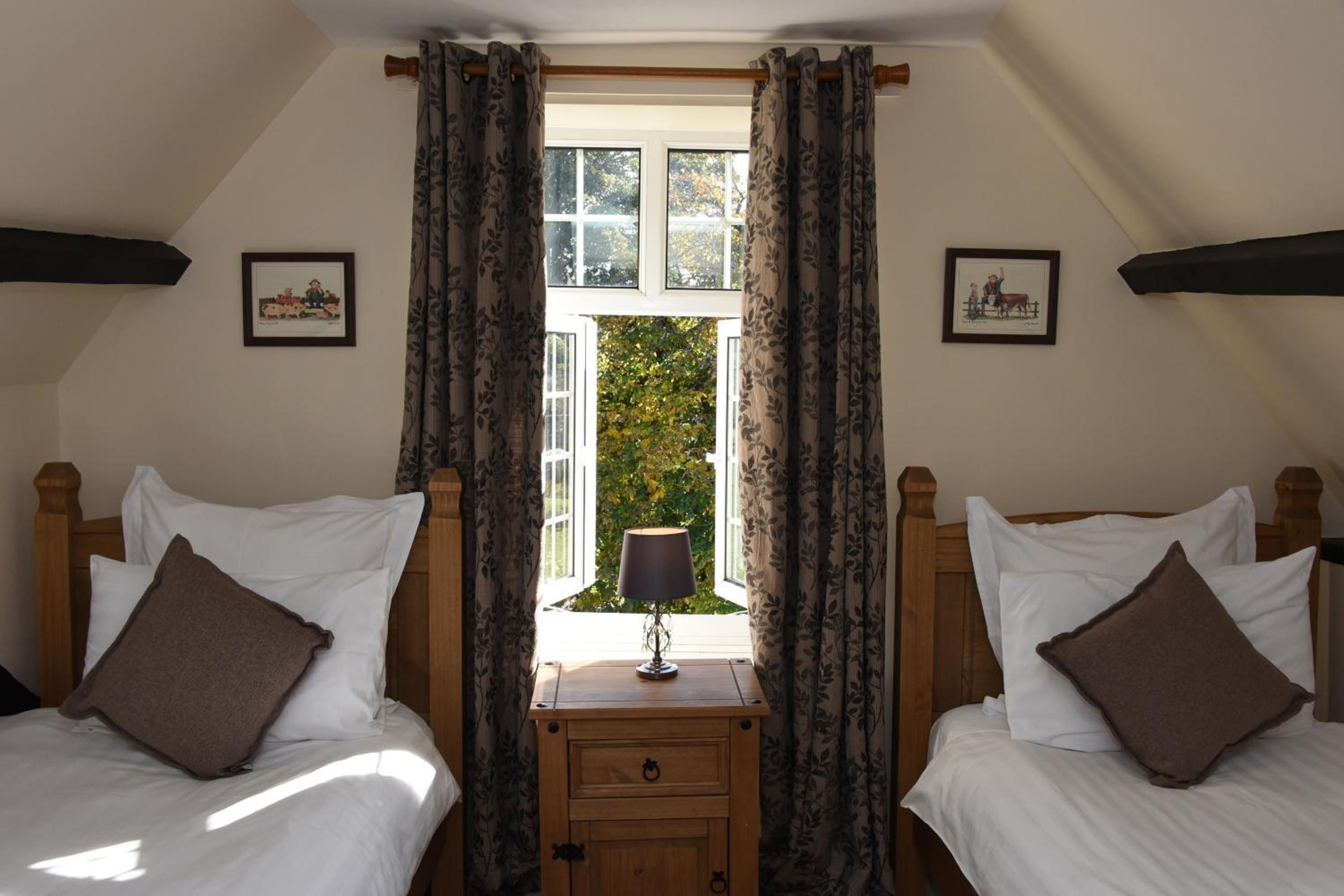 A bedroom with two beds, a window with curtains, a nightstand with a lamp, and two framed pictures on the wall.