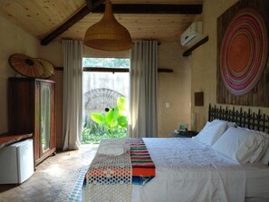 A bedroom with a bed, a small table, a wardrobe, and a view of greenery outside. - Zazen Pousada By Santoregano (Maceió)
