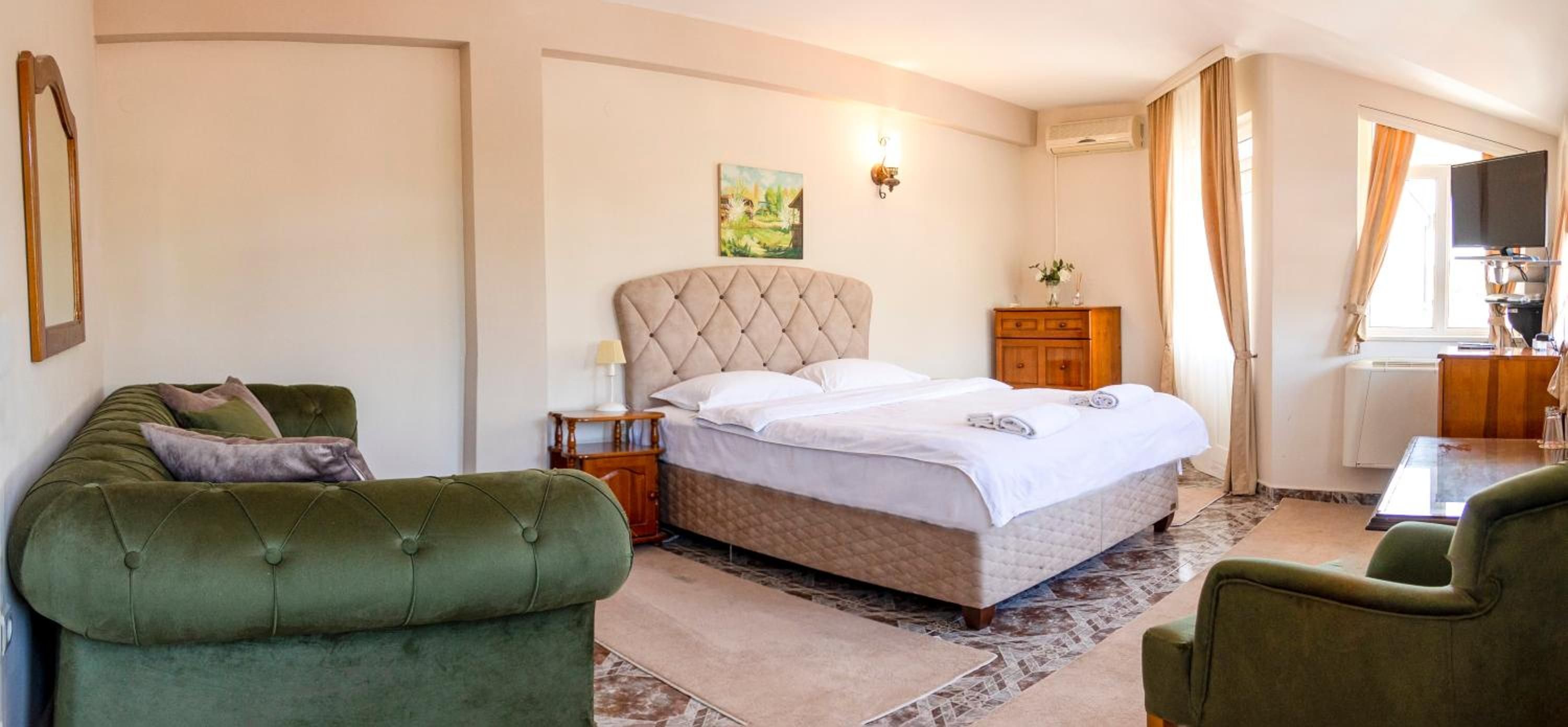 A hotel room with a large bed, two green armchairs, a wooden nightstand, a painting on the wall, and a television.