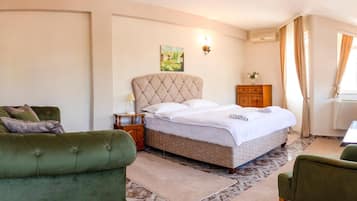 A hotel room with a large bed, two green armchairs, a wooden nightstand, a painting on the wall, and a television.