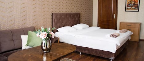 Habitación de hotel con una cama, una mesita de noche de madera, un sofá marrón, una mesa de centro de madera con un jarrón de flores y una alfombra estampada.