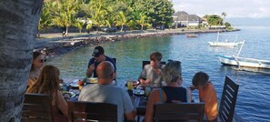 Un grupo de personas cenando en una mesa al aire libre junto a la costa, con un barco amarrado cerca.