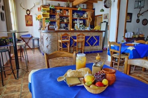 Interior de restaurante con barra, sillas de madera y una mesa dispuesta con pan, fruta y una jarra de jugo.