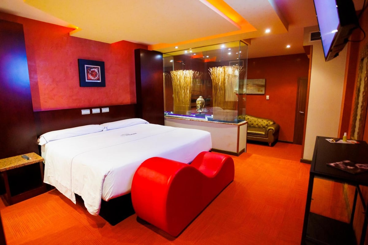 Habitación de hotel con una cama grande, un sillón rojo decorativo, una vitrina con objetos decorativos y un televisor en la pared.