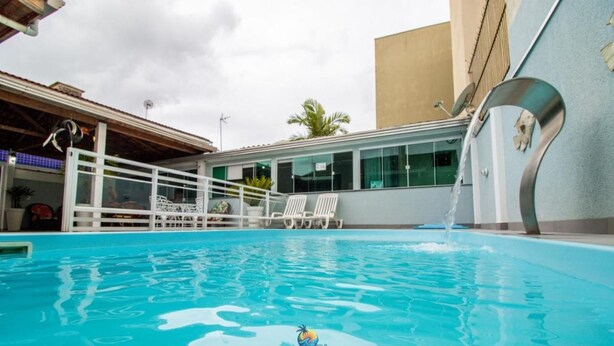 A swimming pool with a water feature, white lounge chairs, and a building with large windows. - Pousada Praia Brava (Matinhos)