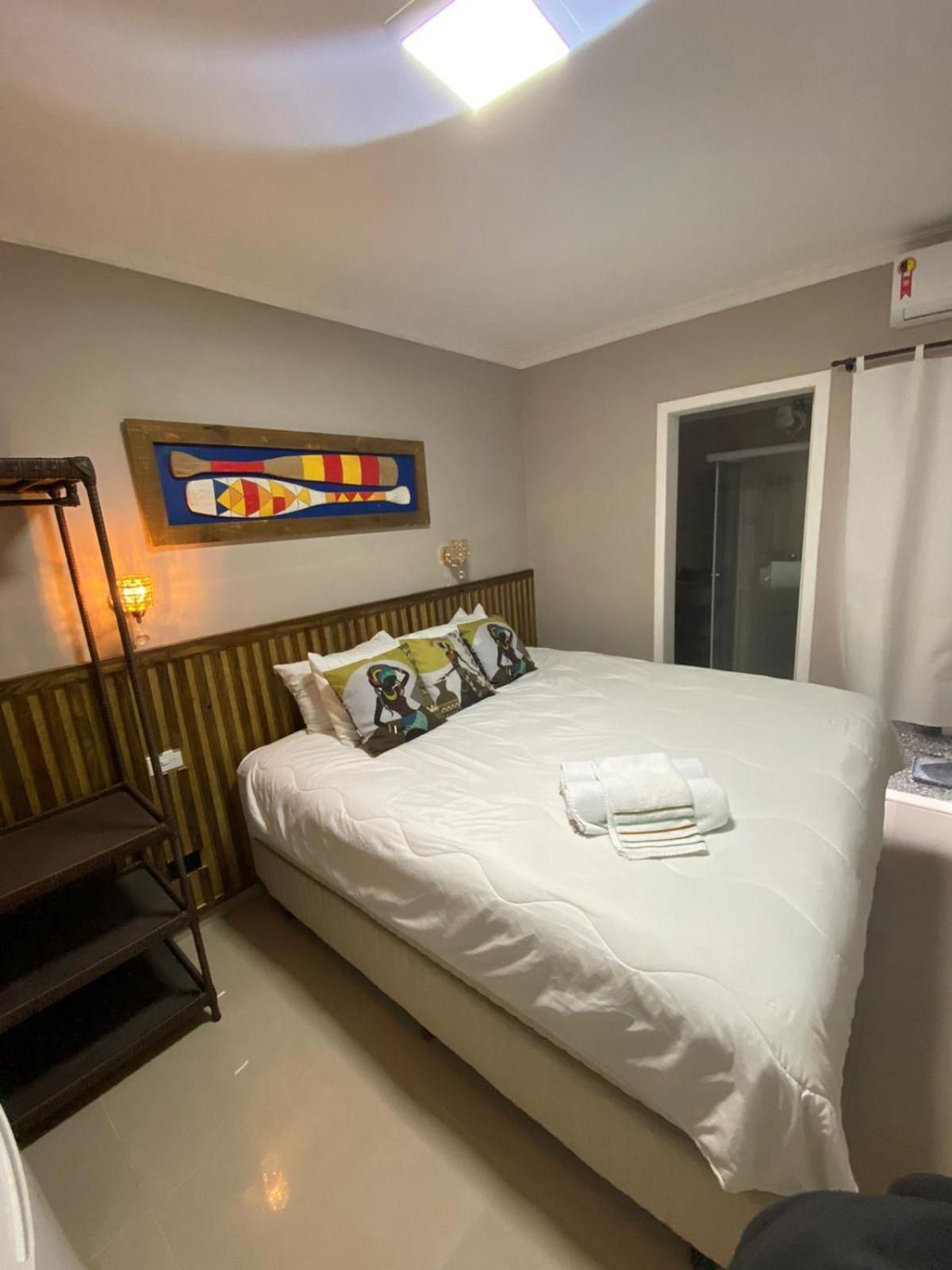 Quarto de hotel com cama, obra de arte na parede, janela com cortinas e mesa de cabeceira.
