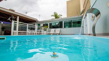 Una piscina con fuente, sillas de descanso blancas y un edificio con ventanas amplias.