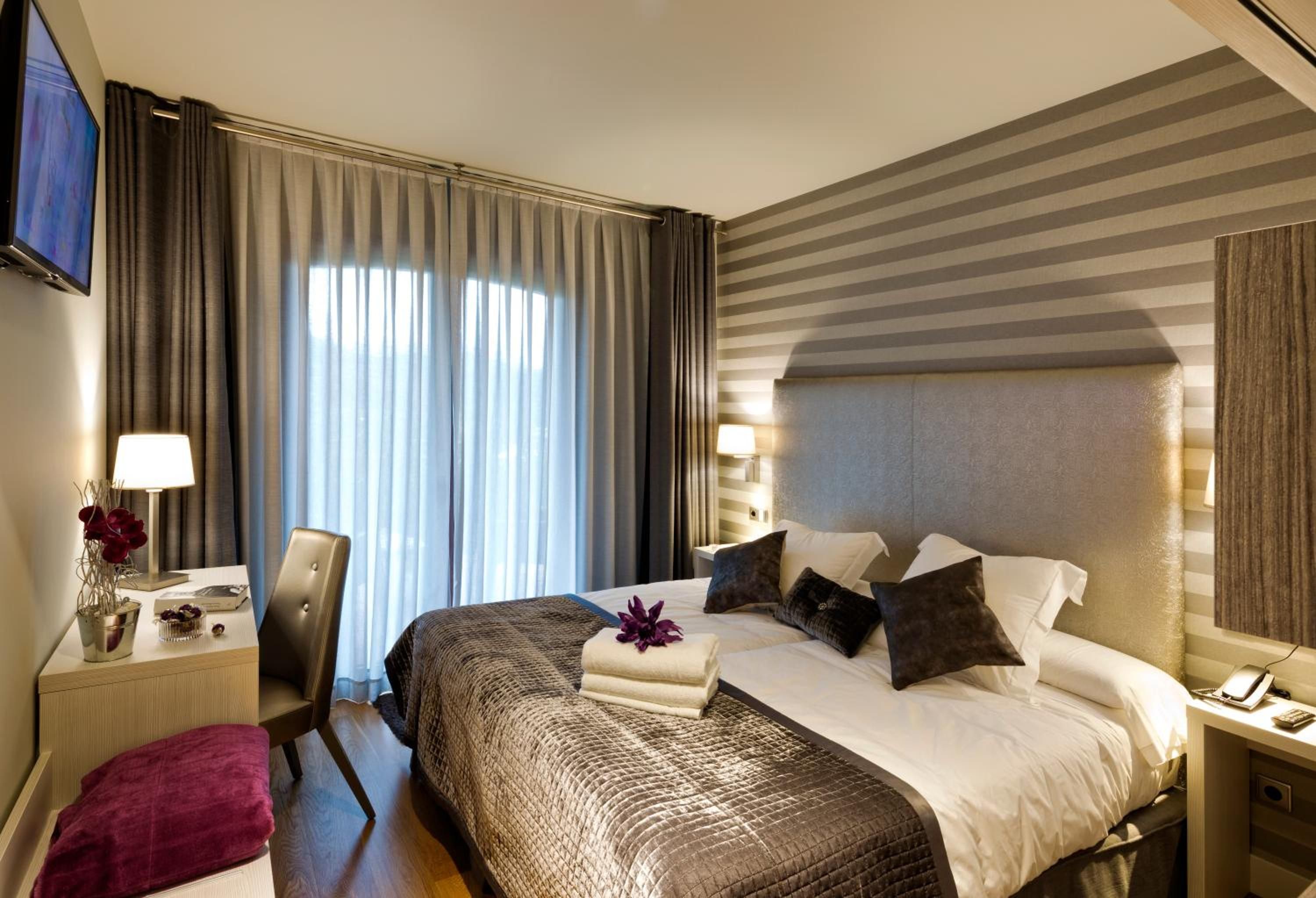 A hotel room with a bed, bedside tables, a desk, a chair, a TV, and a window with curtains.