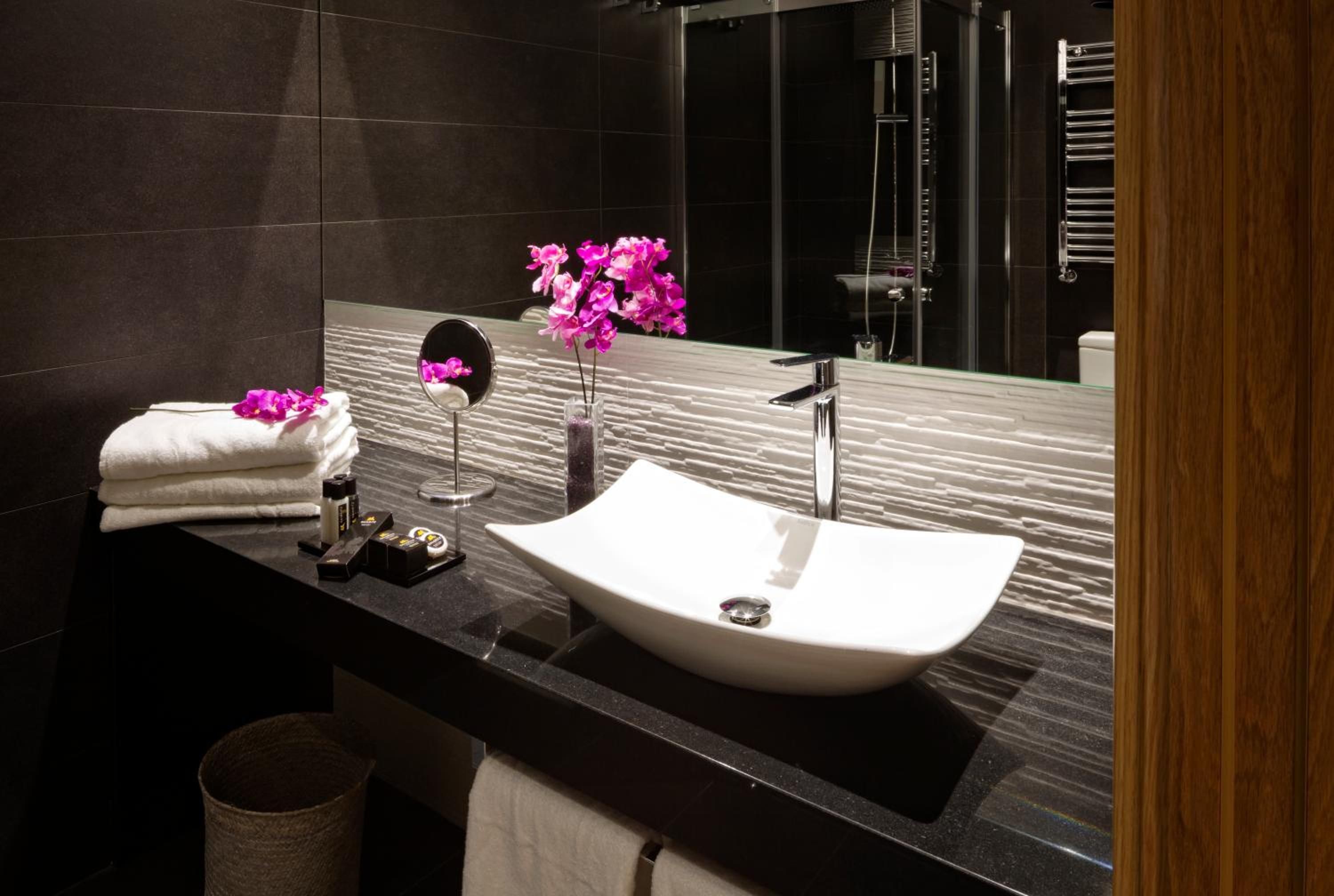 A modern bathroom with a black countertop, a white sink, a mirror, and a vase with pink flowers.