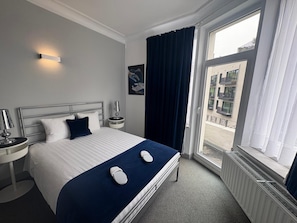 A hotel room with a bed, a nightstand, a lamp, a window with curtains, and a view of the outside. - Eurotel (Liege)
