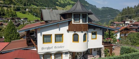 Un edificio de dos pisos con una torre destacada, identificado como 'Berghof Geisler', rodeado por un paisaje montañoso.