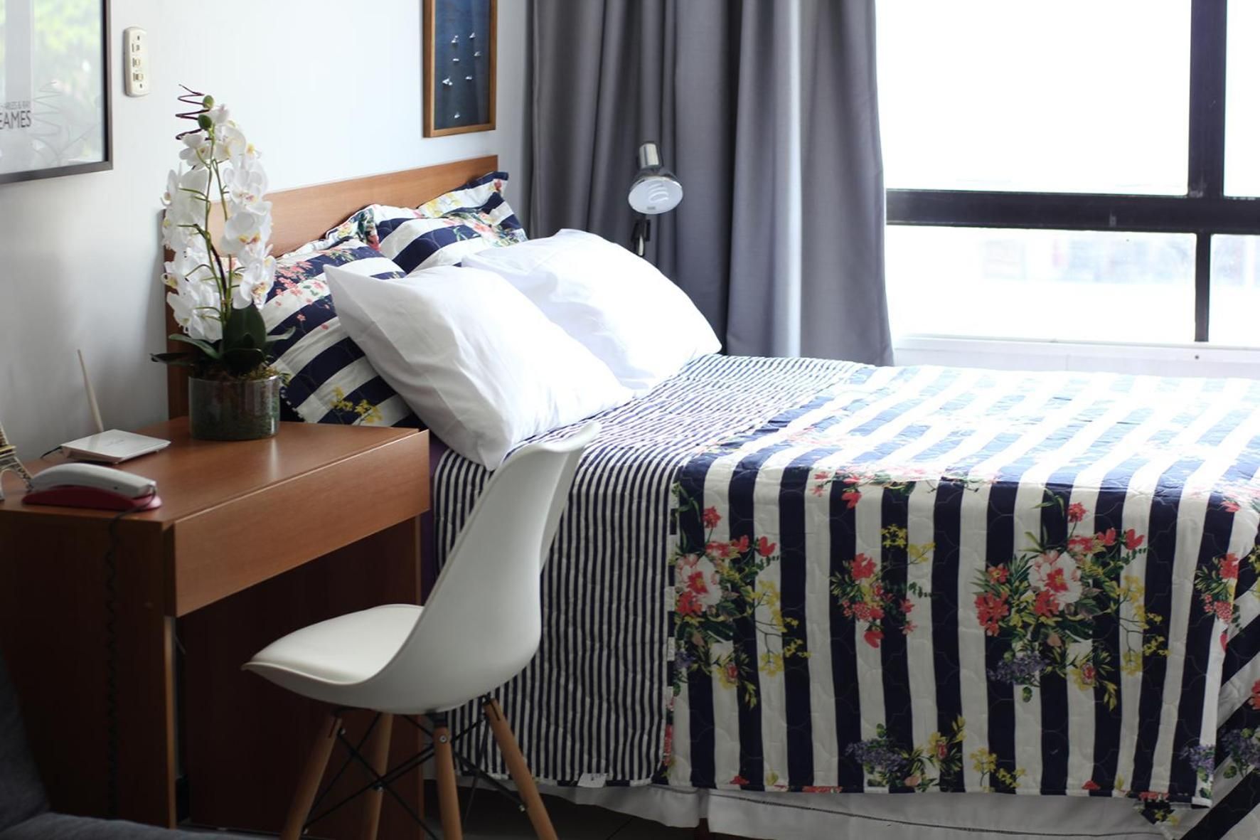A hotel room with a bed, a desk, a chair, a potted plant, and a window with curtains.