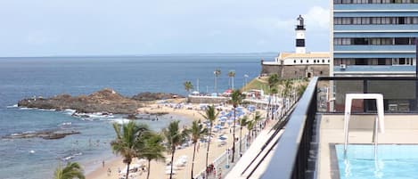 A beach with a pool, a lighthouse, and a building with balconies overlooking the ocean.
