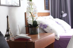 Habitación de hotel ordenada con mesita de noche de madera, una orquídea en maceta pequeña, una réplica de la Torre Eiffel, un teléfono y una cama con colcha de flores moradas y blancas.