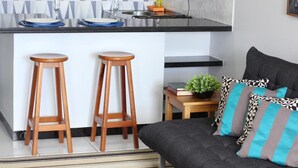 A modern kitchen with a black countertop, two wooden stools, a small table with a plant, and a grey sofa with patterned cushions. - Flat Farol Da Barra (Salvador)