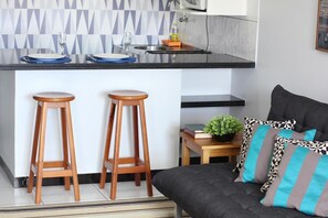 Una cocina moderna con encimera negra, dos taburetes de madera, una mesita con una planta y un sofá gris con cojines estampados.