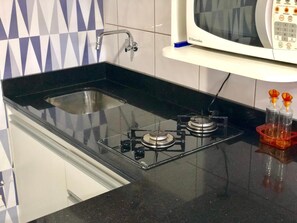 Una cocina con encimera negra, un lavamanos de acero inoxidable y un microondas sobre un estante blanco.