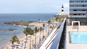A beach with a pool, a lighthouse, and a building with balconies overlooking the ocean. - Flat Farol Da Barra (Salvador)