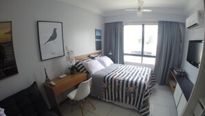 A hotel room with a bed, a desk, a chair, a television, and a window with curtains. - Flat Farol Da Barra (Salvador)