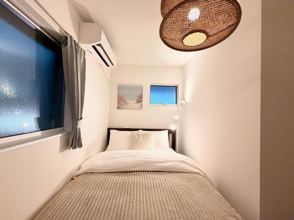 Een slaapkamer met een bed, een raam met gordijnen, een wandairconditioner, een plafondlamp en een klein schilderij aan de muur.