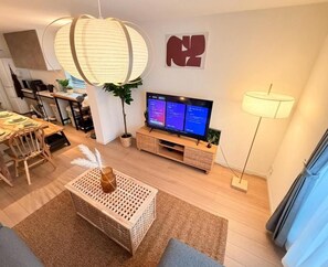 Ein modernes Wohnzimmer mit Fernseher, eine Essecke mit Tisch und Stühlen sowie ein großes Fenster.