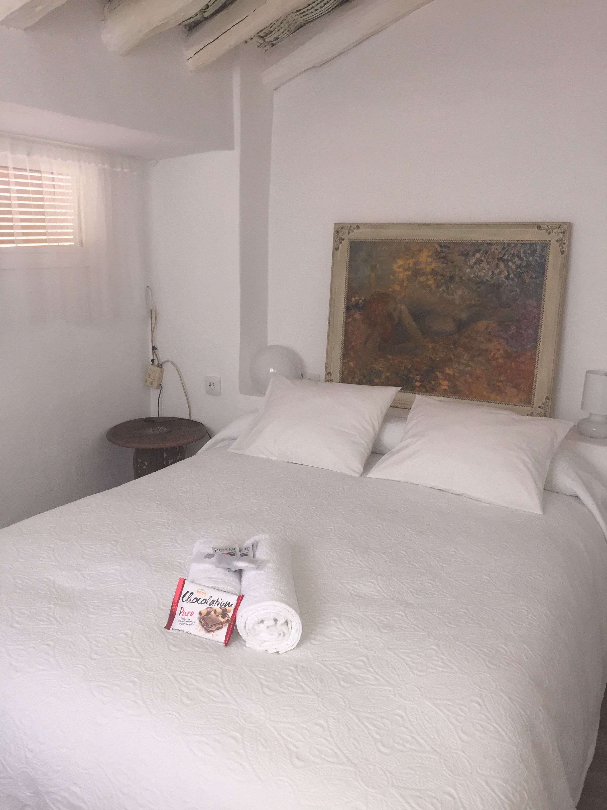 Ein ordentlich bezogenes Bett mit einem gerollten Handtuch und einer Schokoladentafel, ein Gemälde an der Wand und eine Nachttischlampe.