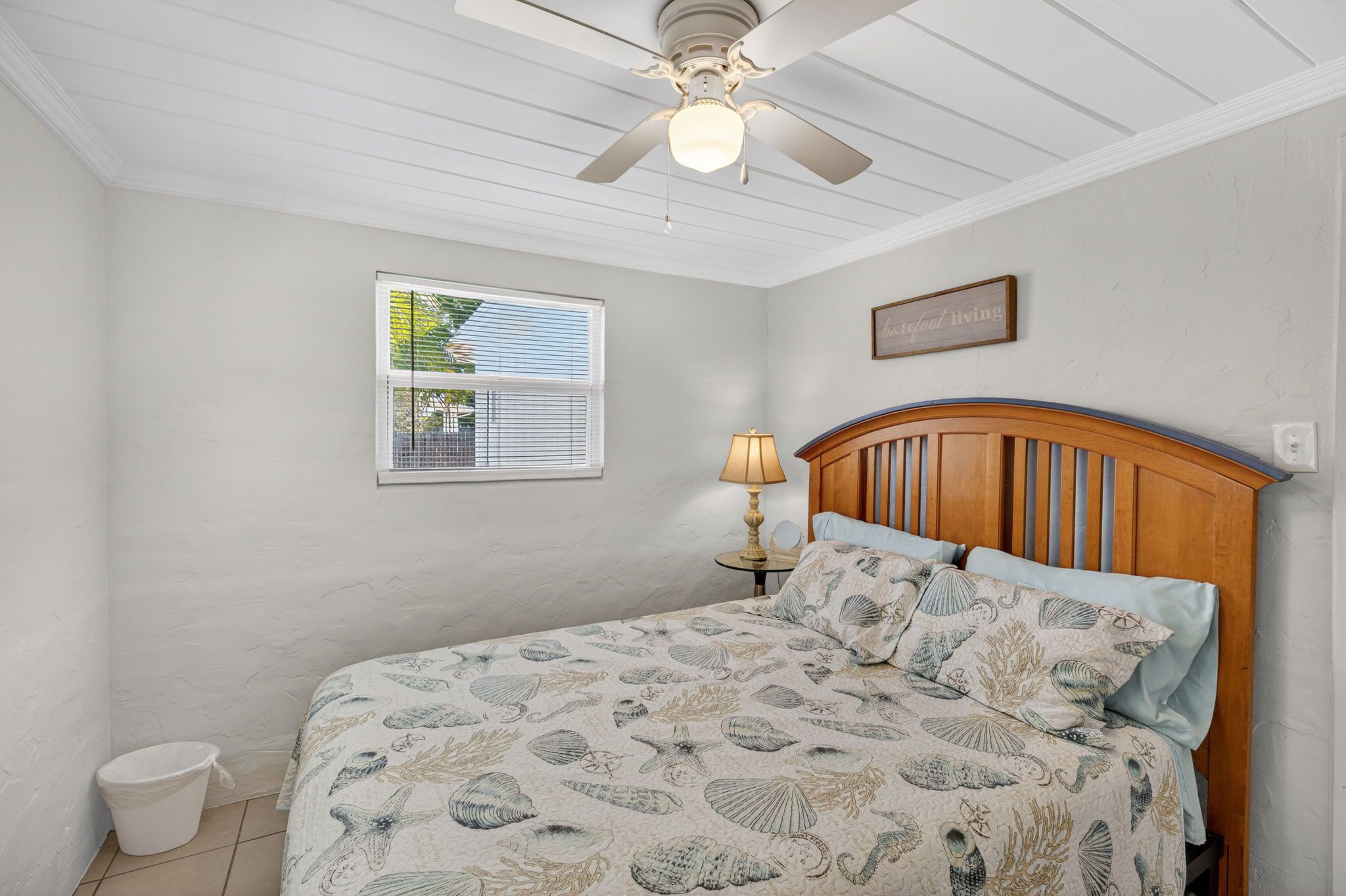 A bedroom with a bed, a ceiling fan, a window with blinds, a bedside lamp, and a wall sign.