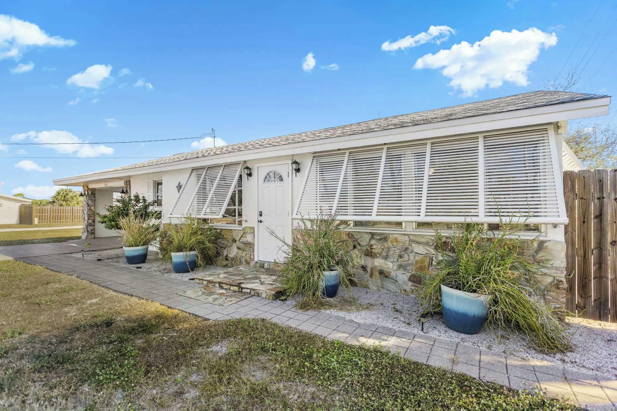 Une maison de plain-pied avec des fondations en pierre, un revêtement extérieur blanc et un toit en tuiles. On peut voir trois grands pots de plantes devant la maison, et une clôture en bois sur la droite.