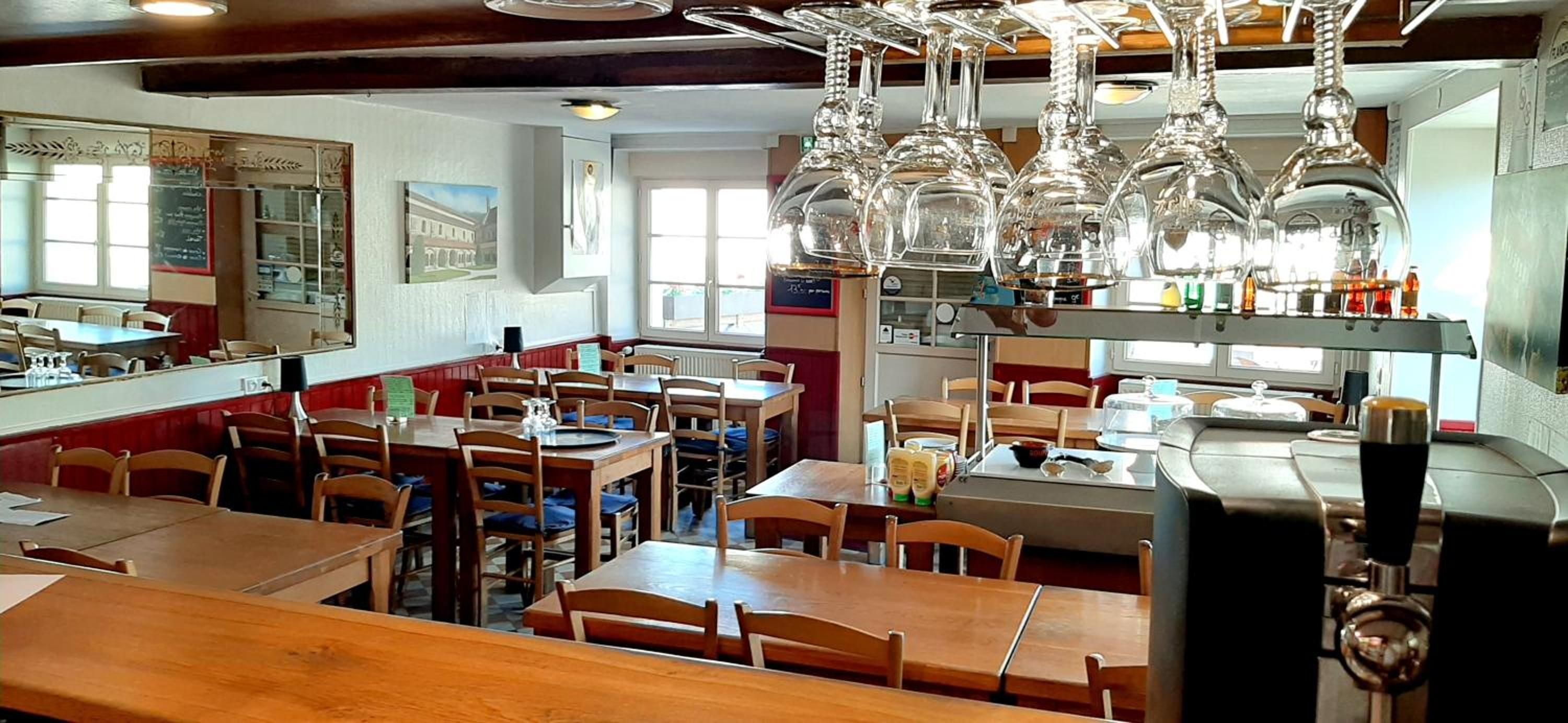 L’intérieur d’un restaurant, avec des tables et des chaises en bois, des luminaires suspendus et un comptoir où sont exposés divers articles.