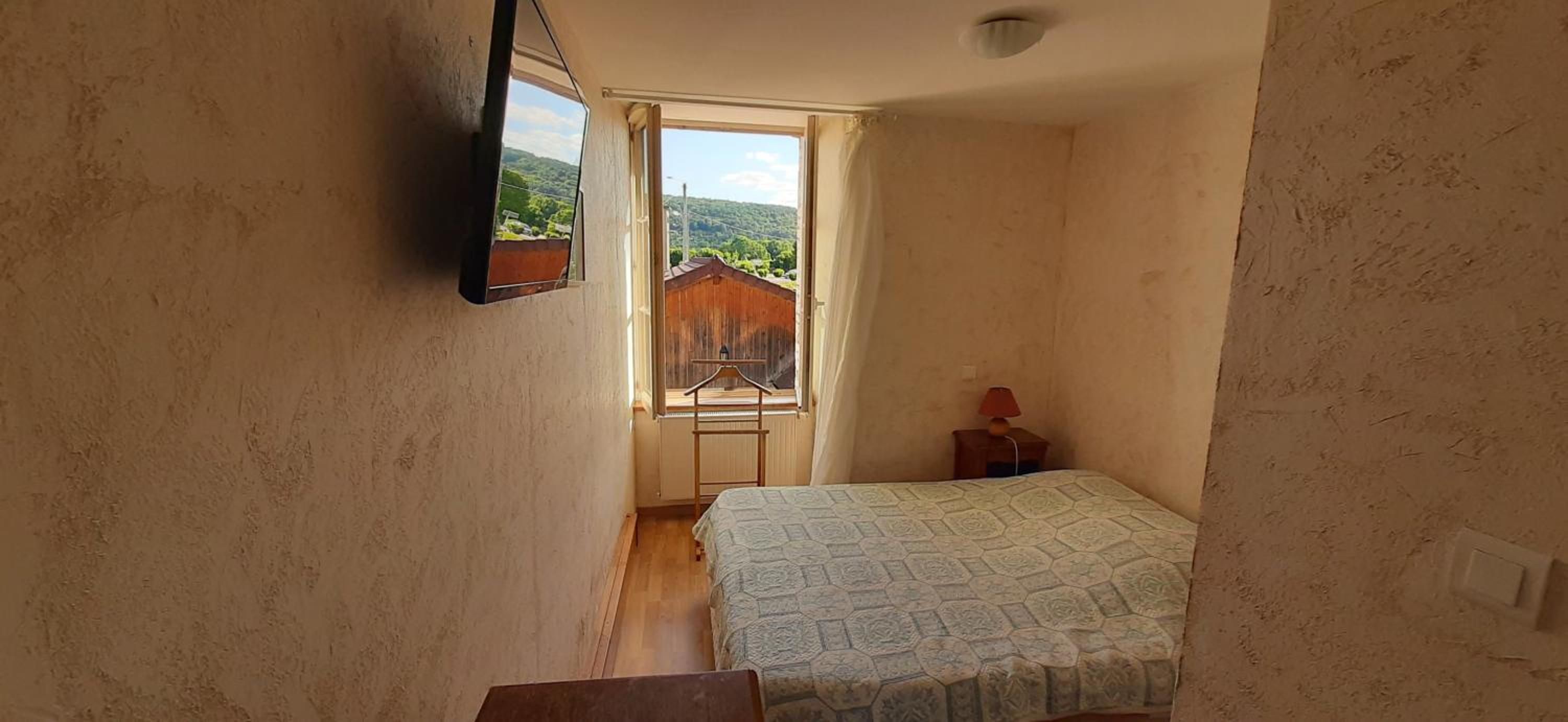 A small bedroom with a bed, a nightstand, a window, and a view of a wooden fence and greenery outside.