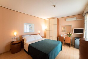 A hotel room with a bed, bedside table, lamp, TV, and a small desk with a chair. - Montebello Park Hotel (Perugia)