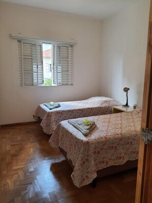 A room with two beds, a window with shutters, a bedside table with a lamp, and a wooden floor. - Pousada Monte Alegre (São Paulo)