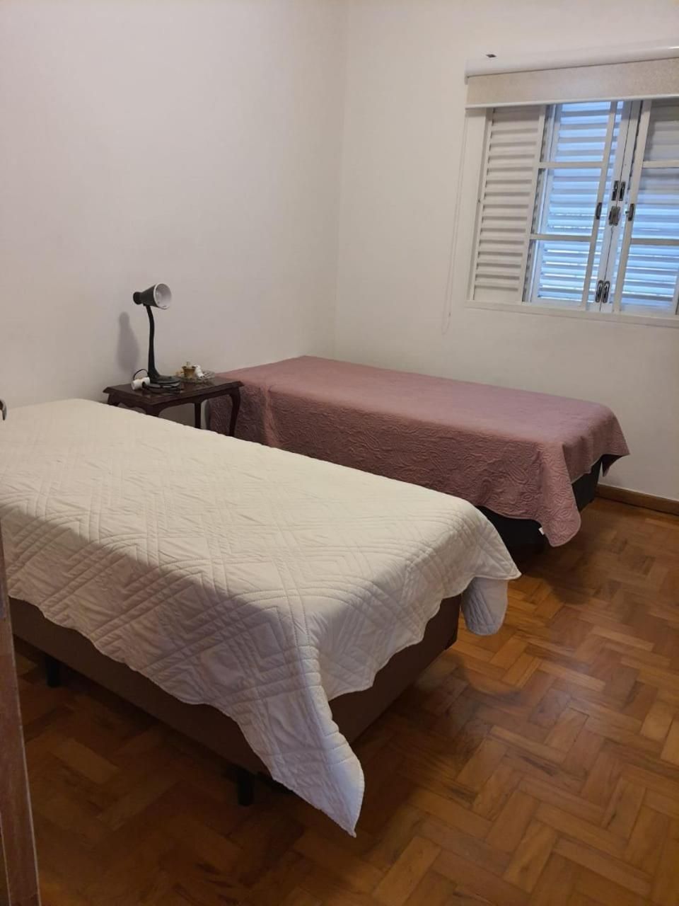 Quarto com duas camas, mesa de cabeceira, abajur e janela com persianas.