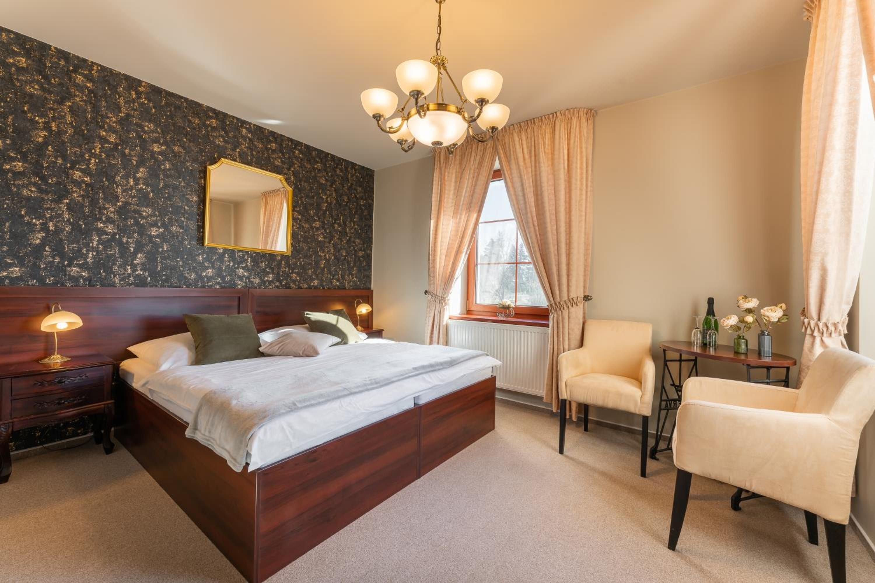 A hotel room with a large bed, a bedside table with a lamp, a chair, a small table with a vase of flowers, and a window with curtains.