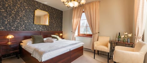 A hotel room with a large bed, a bedside table with a lamp, a chair, a small table with a vase of flowers, and a window with curtains.