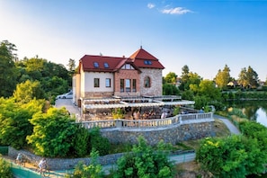 Um edifício de dois andares com fachada de pedra e telhado vermelho, rodeado por vegetação exuberante e um lago.
