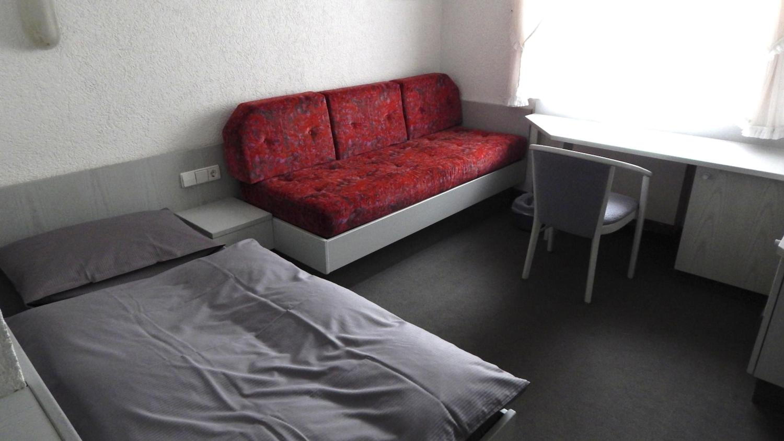 A small, compact room with a bed, a red sofa, a desk, and a chair.