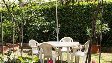 Um alpendre externo com mesa redonda e cadeiras, rodeado por plantas em vasos e um guarda-sol branco.