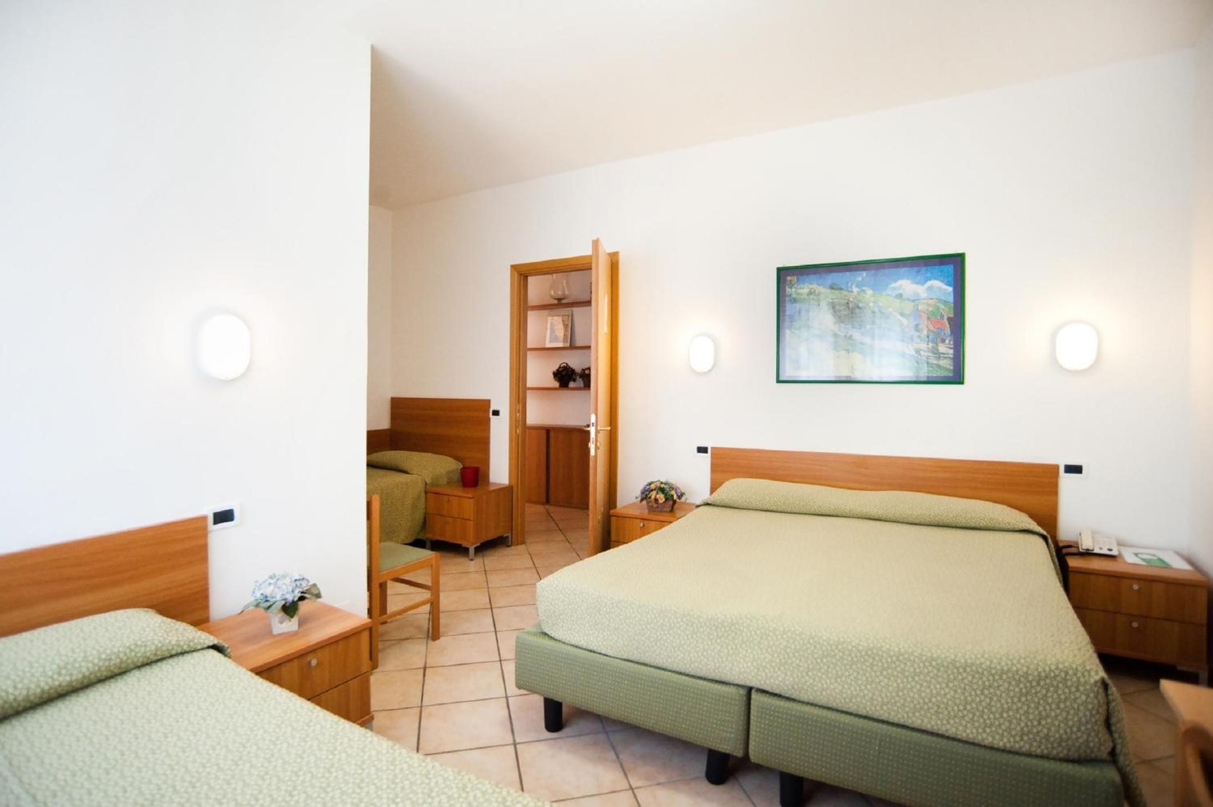 Una camera d'albergo con un letto grande, un letto più piccolo, una scrivania, una sedia, un comodino, un armadio e un quadro appeso al muro.