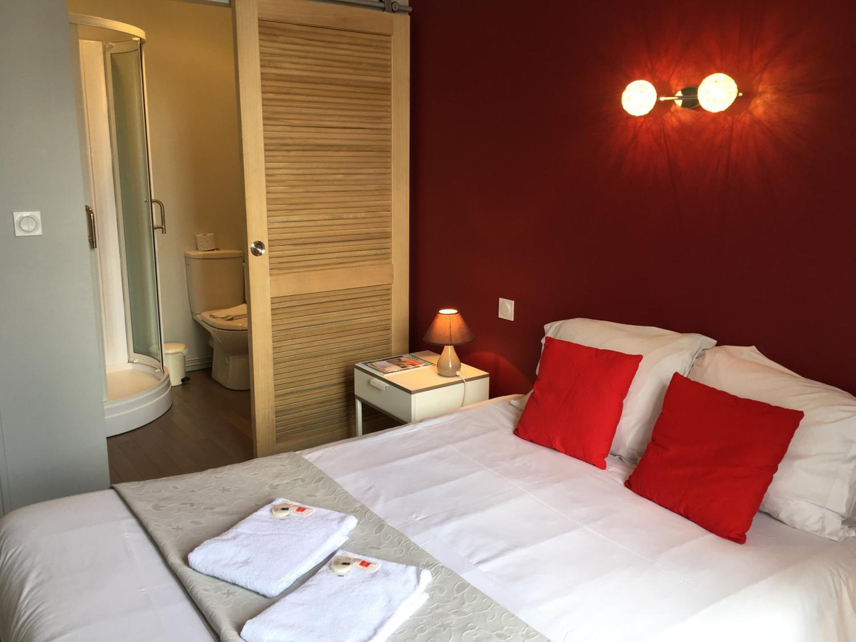 A hotel room with a bed, a nightstand, a lamp, a red wall, and a bathroom with a toilet and shower.