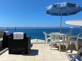 Un balcón con vista al océano, una silla de mimbre negro, una mesa de plástico blanco y una sombrilla azul.
