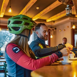 Una persona con casco y chaqueta sentada a una mesa con una taza, en una habitación con vigas de madera y luces colgantes.