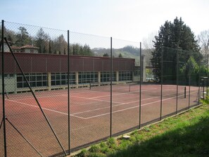A tennis court with a chain-link fence, a building with large windows, and a forested area in the background.